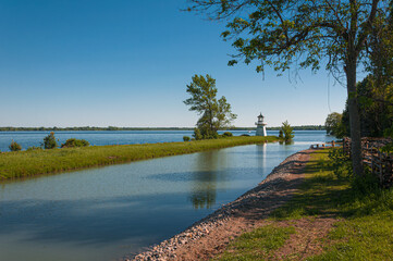 Peaceful summer scene with a white lighthouse on the riverbank, surrounded by lush greenery and blue sky. Calm water reflects the trees and lighthouse, evoking serenity and nature’s beauty.
