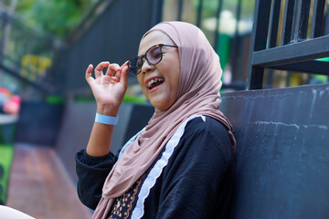 A joyful asian woman in a hijab laughs while adjusting her sunglasses, enjoying a relaxing moment
