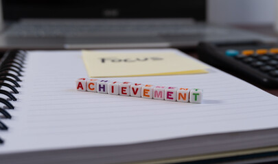 A stack of colorful cubes with the word "achievement" written on them. The cubes are arranged in a row on a notebook, with a sticky note reading "focus."