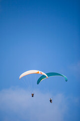 A pair of paragliding athletes are floating in the air showing their skills in controlling their paraglider against a backdrop of bright blue sky.