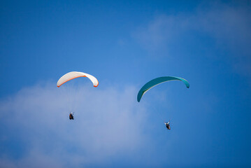 A pair of paragliding athletes are floating in the air showing their skills in controlling their paraglider against a backdrop of bright blue sky.