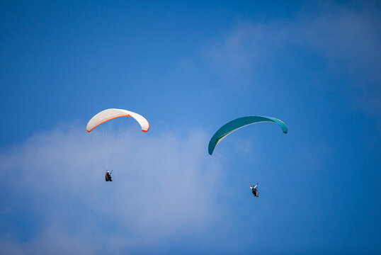 A pair of paragliding athletes are floating in the air showing their skills in controlling their paraglider against a backdrop of bright blue sky.