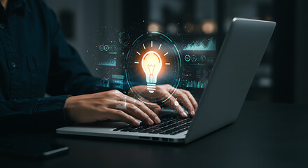 Fototapeta premium Creative Innovation: Hands type on a laptop with glowing light bulb graphic, representing new ideas and innovative solutions.