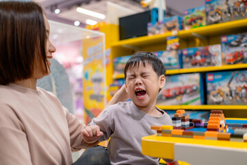 Stubborn child refusing to leave toy store while building with colorful bricks, making mother tired