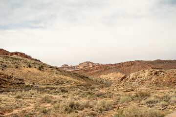 A rugged desert canyon scene with sparse brush, rocky outcrops, and sandstone cliffs under a cloudy sky in the American Southwest