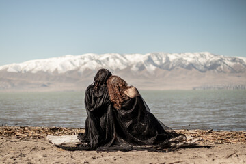 Two women draped in black cloaks sit close together on a sandy lakeshore, facing a snow-covered mountain range in the distance
