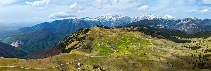 Panoramic aerial view of green highland meadows with mountain huts and snow-capped peaks in the...
