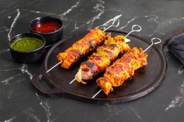 Grilled chicken skewers on a black plate with sliced tomatoes, onions, lime, green chutney, and ketchup, served on a dark marble background.
