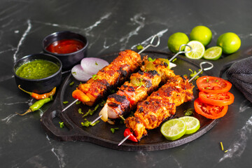 Grilled chicken skewers or chicken tikka kebab on a black plate with sliced tomatoes, onions, lime, green chutney, and ketchup, served on a dark marble background.
