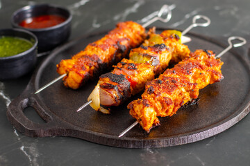 Grilled chicken skewers on a black plate with sliced tomatoes, onions, lime, green chutney, and ketchup, served on a dark marble background.
