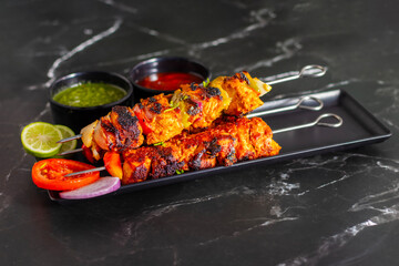 Grilled chicken skewers or chicken tikka kebab on a black plate with sliced tomatoes, onions, lime, green chutney, and ketchup, served on a dark marble background.
