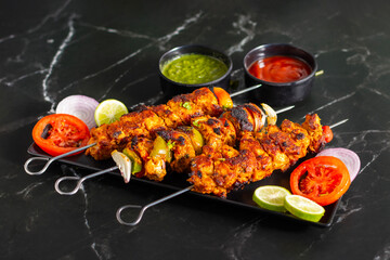 Grilled chicken skewers or chicken tikka kebab on a black plate with sliced tomatoes, onions, lime, green chutney, and ketchup, served on a dark marble background.
