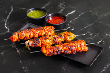 Grilled chicken skewers on a black plate with sliced tomatoes, onions, lime, green chutney, and ketchup, served on a dark marble background.
