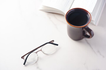 白いテーブルに置かれた一冊の本とメガネ、コーヒーの入ったマグカップで、コーヒーを飲みながら読書を楽しんでいるイメージ
