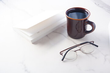 白いテーブルに置かれた一冊の本とメガネ、コーヒーの入ったマグカップで、コーヒーを飲みながら読書を楽しんでいるイメージ
