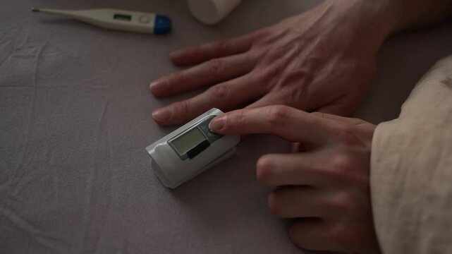 Patient uses a pulse oximeter on their finger to check oxygen saturation levels, with a digital thermometer visible in the background on a bedsheet