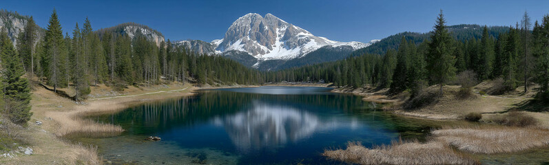 Panoramic mountain lake reflection: A breathtaking landscape captures the serene beauty of a mountain lake, reflecting snow-capped peaks and lush forests, all under a clear blue sky. 