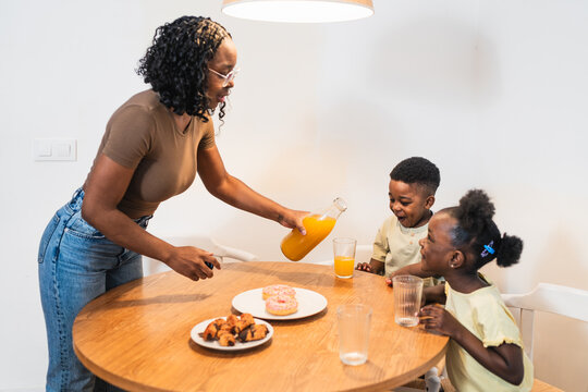 Happy african family enjoying breakfast together at home, mother pouring orange juice for her two children