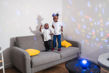 African two cheerful children are jumping and dancing on a sofa in the living room, enjoying a playful moment at home