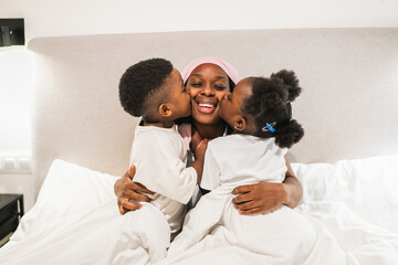 Two children kissing their smiling mother wearing a pink headscarf, lying in bed and fighting cancer