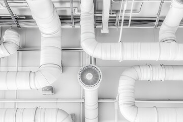 White industrial ventilation and ductwork system overhead.  Complex network of pipes and ducts crisscrossing a ceiling.  A circular fan is centrally located