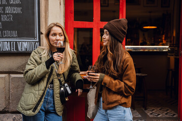 Side view of a dark-haired European woman in her early 20s wearing a brown hat, holding a glass of wine, and watching as her blonde young female friend tastes wine and talks at a bar.