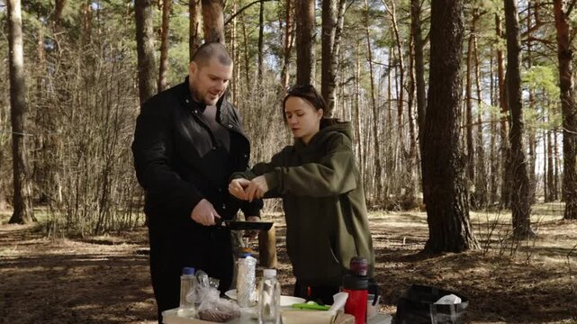 Two people preparing a meal outdoors in a pine forest, with one person cracking eggs into a frying pan held over a compact camping stove