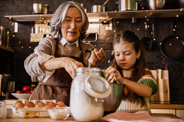 A White grandmother in her 60s with gray hair and a school-aged granddaughter standing at a table with eggs and flour while they are kneading the dough in a metal bowl in a kitchen with black walls