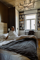 Waking up in the morning in a hotel room, a sad dark-haired European man in his early 40s dressed in a white T-shirt sits on the bed, covered in a gray blanket.