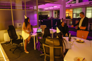 In the office, a gray-haired European director in his early 50s congratulates his dark-haired 30-year-old colleague on his birthday while other multi-ethnic adult colleagues look on in the office.