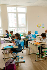 In the foreground, a serious dark-haired European school-aged girl sits at a desk while other multi-ethnic schoolchildren draw in the classroom during the daytime.