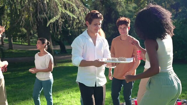 Young multiethnic friends sharing food, laughing while enjoying sunny picnic in green park, capturing carefree summer leisure moment through slow motion