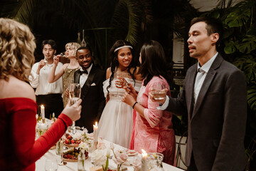 A couple in their 30s in wedding attires standing at a festive table with candles, food, and drinks in a hall with a multiethnic group of adult wedding guests with glasses of wine during a celebration