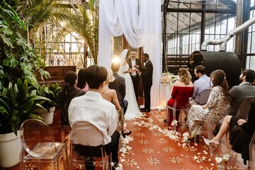 At a restaurant at a wedding ceremony, a European adult master of ceremonies and multi-ethnic guests watch as a Black 30-year-old groom puts a ring on his 20-year-old bride in a white dress.