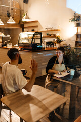 A female waitress in an apron working at a laptop while sitting at a table in a cafe and talking...