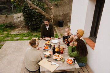 A multiethnic group of adult friends gathered around a table with plates of vegetables, fruits, bread, and red wine and talking in a cozy yard in front of a white house and trees around them