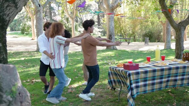 Group of diverse friends forming a conga line and dancing at a picnic in a park, having fun in slow motion