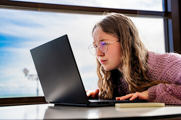 Young woman with glasses squinting at laptop screen while working indoors at daylight, female dont...