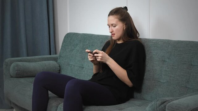 Young woman relaxing at home, carefully filing and shaping natural nails while sitting on comfortable gray sofa, showcasing personal beauty and wellness routine