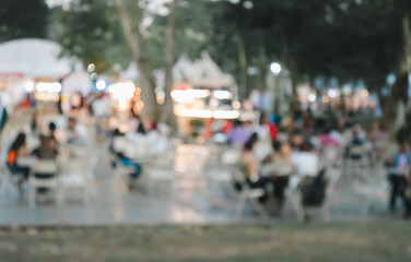 Blurred image of outdoor gathering with people sitting at tables under trees. Night festival market in green park 