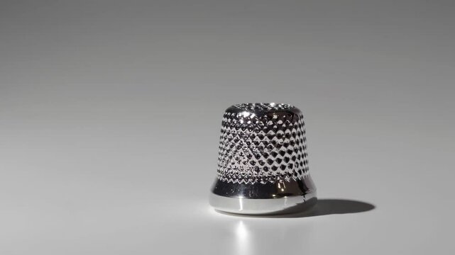 Precision metallic thimble rotating slowly, casting intricate light and shadow patterns against pristine white background