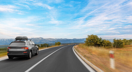 A black SUV with a roofbox drives along a winding country road surrounded by trees and mountains under a wide, clear blue sky.