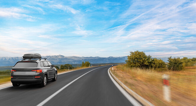 A black SUV with a roofbox drives along a winding country road surrounded by trees and mountains under a wide, clear blue sky.