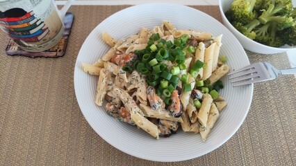 Penne pasta with creamy dill sauce, mussels, and chopped spring onions, served on a white plate with a fork and a side of broccoli