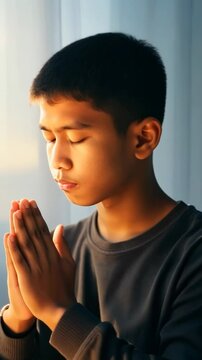 Young man praying with hands joined in meditation pose against soft light background. His expression changes subtly from concentration to peaceful serenity as sunlight creates warm atmospheric glow.