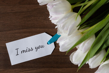 I miss you message tag placed near white tulips on wood, expressing feelings of emotion, longing,...