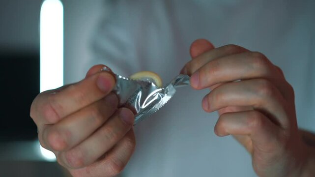 Condom package in hands.
Man opens condom.
Protection from viruses during sex.
Safe sex.
Sexual education.
Shiny condom package
