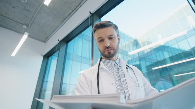 Close up of Caucasian male doctor reading medical reports near large window in bright clinic, reviewing patient information while standing in modern workspace. Review of analysis in hospital.