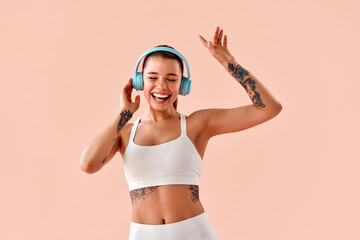 Stylish young woman with arm tattoos, wearing a white sports top and leggings, dancing with blue headphones, enjoying a fitness workout and maintaining a positive mood, against a soft pink background.