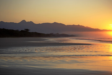 Sunrise in Itaguare beach, Bertioga, Brazil. Yellow light on sky and reflections on water, shadows over vegetation in silhouette and  mountains in background